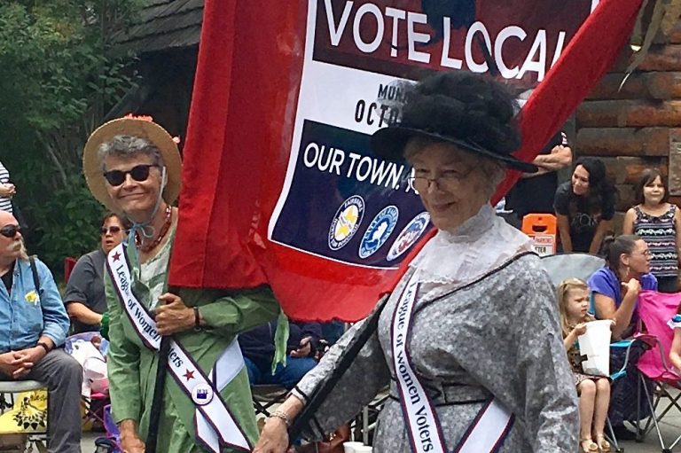Image of two women dressed as suffragists carrying a banner in a parade.in a parade
